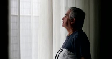 Pensive older man standing by window looking outside