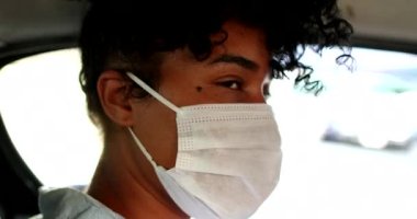 Salgın yüz maskesi takan genç siyahi kadın taksinin arka koltuğunda kameraya bakıyor.