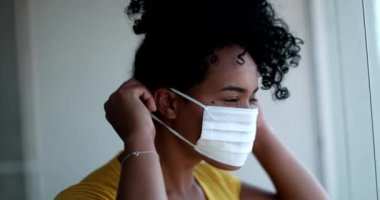 Young African American woman putting virus prevention mask, black girl standing by window looking outside