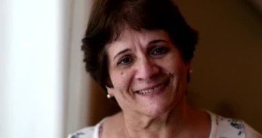 Casual older 60 year old woman smiling at camera standing by window, portrait face