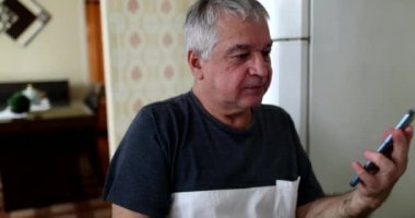 Older man putting reading glasses checking smartphone device at home