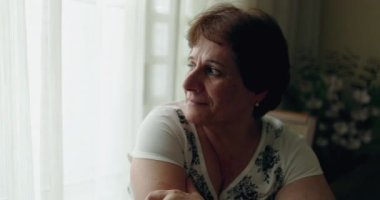 Pensive older senior woman sitting at home by window