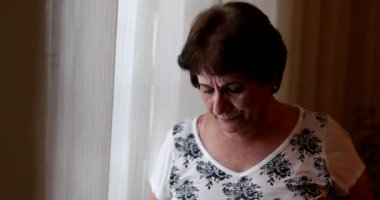 Sad older woman standing by window at home. Depressed worried 60 year old lady