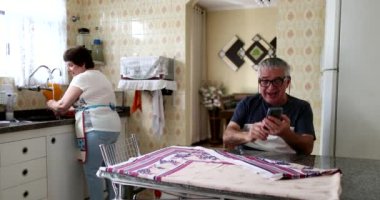 Older married couple receiving good news on smartphone. Senior sitting at kitchen commemorates news with wife