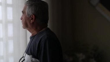 Pensive senior man standing by window putting glasses