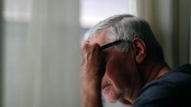 Senior man standing by window looking outside. Elder suspicious person observing neighborhood