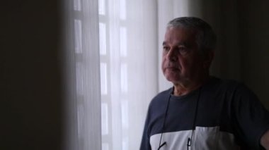 Pensive senior man standing by window putting glasses
