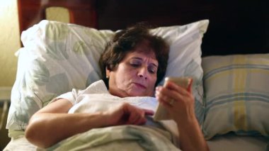 Older woman lying in bed browsing internet on smartphone before sleep