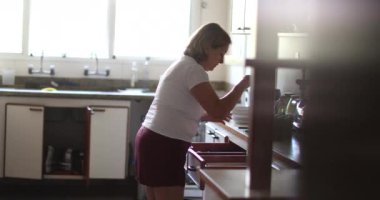 Açık yürekli, yaşlı bir kadın mutfak temizliği yapıyor. Çekmecelerin içinde mutfak gereçleri düzenliyor.