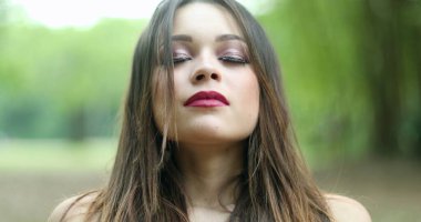 20 'li yaşlarda derin bir nefes alan bir kız. Parkta temiz hava soluyan genç bir kadın kameraya bakıyor kendini yenilenmiş ve mutlu hissediyor.