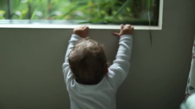 Infant one year old baby standing by window looking outside. Adorable toddler child leaning on window