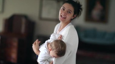 Mother holding baby boy infant toddler in arms at home, adorable child biting clothing casual and authentic