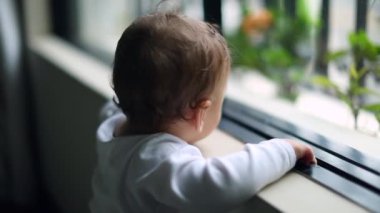 One year old baby standing by home window looking outside