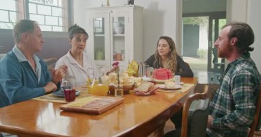 Orta yaşlı ebeveynler, küçük kızı ve büyük oğluyla kahvaltı yapıyor. Anne kocasına peynir ekmeği servis eder. Oğlu ekmek yiyor, mutlu mesut sohbet ediyorlar..