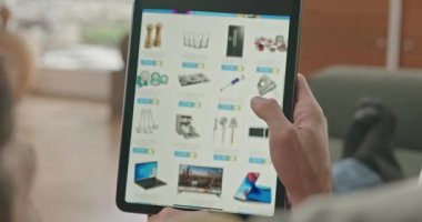 Yakından bakılan eller online alışveriş sitesinde satın alınacak ürünü arıyor. Evdeki koltukta oturan kişi e-ticaret sitesi uygulamasına bakıyor.