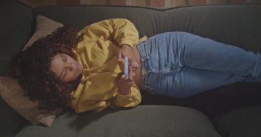One black hispanic girl texting message on cellphone device. From above angle of young woman using phone text messages friend. Person lying on couch sofa using modern technology device