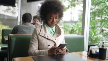 Bir siyah kadın kahve masasında oturan akıllı telefon cihazını kontrol ediyor. Telefonla internette gezinen Afrikalı Amerikalı bir kız.