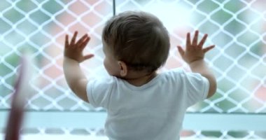 Cute baby leaning on window looking outside. Toddler crawlling on floor