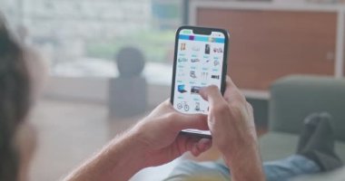 Adam internetteki bir alışveriş ve perakende sitesinde cep telefonuyla internette geziniyor. Oturma odasındaki rahat kanepesinde oturuyor.