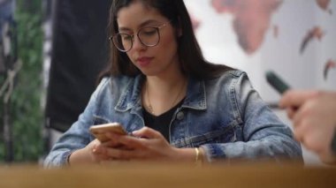 Kahve dükkanında oturan genç ve dürüst bir kadın. 20 'li yaşlarda Güney Amerikalı bir kız akıllı telefon cihazına bakıyor.