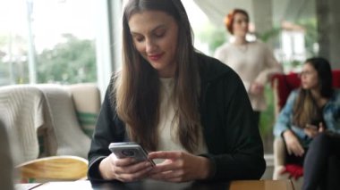 Kafe restoranında oturan akıllı telefondan mesaj okuyan gülümseyen bir genç kadın.