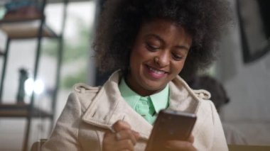 Kahve dükkanında telefonla gülümseyen mutlu bir zenci kadın. Afro-Amerikalı neşeli bir ifadeyle akıllı telefona bakıyor.