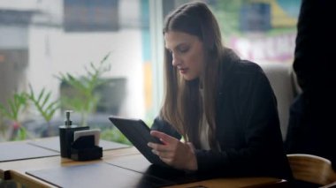 Kafede oturup tablet cihazından içeriği okuyan samimi genç bir kadın. Modern teknolojiyi kullanarak ekrana dikkat eden kişi