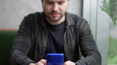 Genç adam cep telefonuyla internetten içeriği okuyor. Kahve dükkanında pencere kenarında oturuyor. Akıllı telefon mesajı kullanan ciddi ifadeli bir kişi