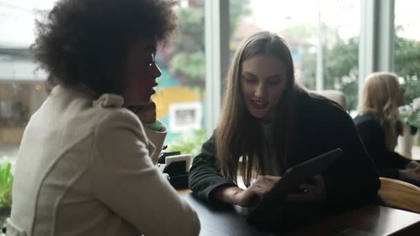 コーヒーショップで女性の友人にタブレット画面を示す若い女性 現代のテクノロジーを見ながらカフェテーブルレストランに座っている二人の女性 — ストック動画