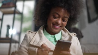 Kahve dükkanında telefonla gülümseyen mutlu bir zenci kadın. Afro-Amerikalı neşeli bir ifadeyle akıllı telefona bakıyor.