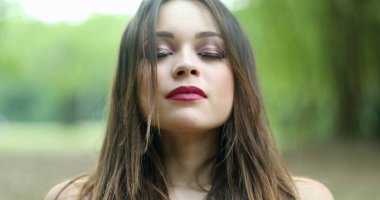 20 'li yaşlarda derin bir nefes alan bir kız. Parkta temiz hava soluyan genç bir kadın kameraya bakıyor kendini yenilenmiş ve mutlu hissediyor.