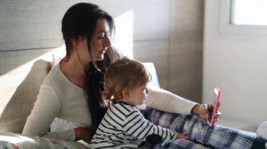 Mother and baby child together in bed holding smartphone. Infant boy and parent in morning taking selfie