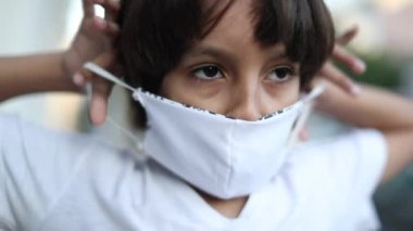 Child putting on covid preventive mask against pandemic outside. Mixed ethnically diverse young boy kid puts surgical mask
