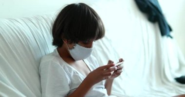 Child wearing face mask and playing video-game at home. Youn boy holding smartphone playing game during pandemic