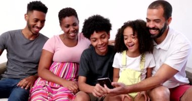Akıllı telefon tutan mutlu Afrikalı ırklar arası aile. Ebeveynler ve çocuklar evdeki koltukta cep telefonuna bakıyor.