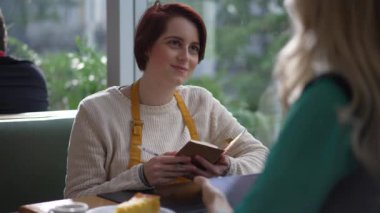 Young female staff listening to manager speak. Girl writing notes for new job sitting at table speaking with boss. Person wearing apron and holding paper and pen