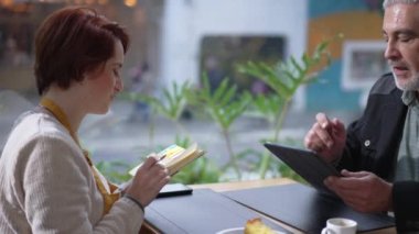 Candid businessman manager showing tablet to female staff seated at coffee shop. Entrepreneur explaining job to new employee. Woman writing notes learning instructions