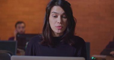 Bilgisayarın önünde konuşan kulaklık takan kadın bir ofis çalışanı. Genç bir kadın, iş yerindeki bilgisayar ekranının önünde gece vakti iş arkadaşıyla video üzerinden konuşuyor.