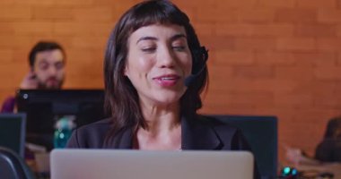 Ofis çağrı merkezinde bilgisayar ekranının önünde kulaklıkla konuşan kendinden emin genç bir kadın. Müşteri hizmetlerinde çalışan bayan çalışan müşteriyle çevrimiçi konuşuyor. Güney Amerikalı çalışanlar gece çalışma alanında