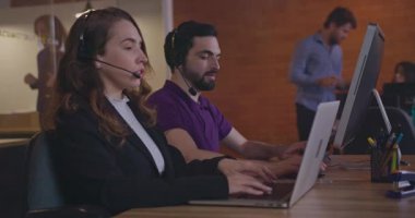 Tele pazarlamacılar dizüstü bilgisayarların önünde kulaklık takan müşterilerle iletişim kuruyor. Ofis içinde çalışan kadın ve erkek çalışanlar çağrı merkezindeki müşterilerle konuşuyor.