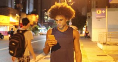 African mixed race young man holding cellphone device looking at screen while walking at night in downtown city during drizzle