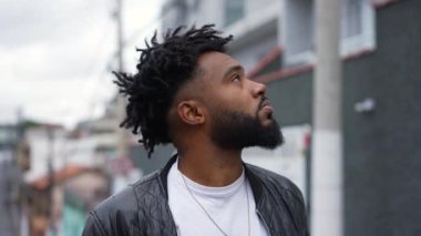 A pensive African American man looking up to sky in contemplation standing by urban street