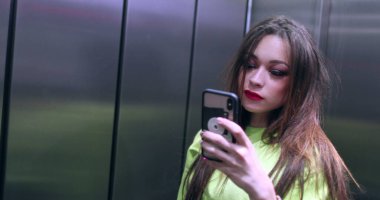 20 'li yaşlarda güzel bir kız asansörde aynanın önünde telefonla fotoğraf çekiyor. Çekici genç kadın selfie çekiyor.