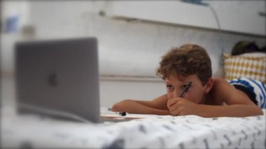 Young boy studying at home in front of laptop. Child doing homework lying in bed in front of computer