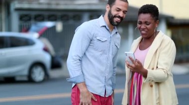 Couple laughing at video on cellphone outside in street. Mix race married couple authentic laugh and smile looking at smartphone