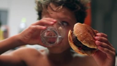 Young boy eating meal. Kid drinking water and taking a bite of hamburger