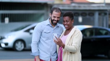 Mix race married couple laughing holding smartphone in street, authentic real life laugh and smile