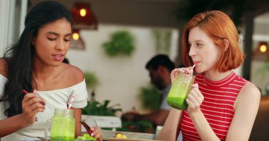 Çekici iki genç çok ırklı kadın yemek yiyor ve taze yeşil smoothie içiyor.