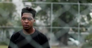 Angry young black man looking to camera leaning on metal fence. Upset African American