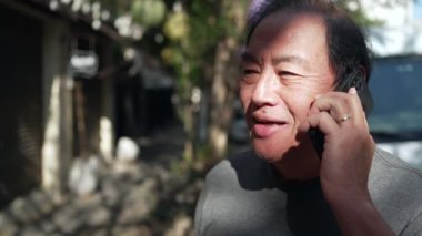 A happy middle aged Asian man speaking on cellphone standing outdoors in street. Person holding smartphone in ear in conversation